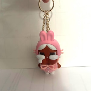 Puede incluir: Un llavero rosa con temática de conejito con un anillo y una cadena dorados. El llavero presenta una pequeña figura de piel morena que lleva una capucha y un atuendo de conejito rosa, con detalles blancos y un lazo. La figura tiene una expresión triste con lágrimas blancas.
