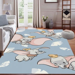 May include: A rectangular area rug featuring a repeating pattern of the Disney character Dumbo the elephant in flight. The rug has a light blue background with white feather accents and Dumbo illustrations in gray, pink, and yellow.