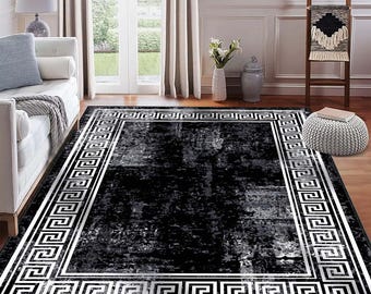 May include: A rectangular black and gray area rug with a white Greek key border. The rug has a distressed, textured appearance, adding a touch of elegance to the room. The rug is placed on a hardwood floor.