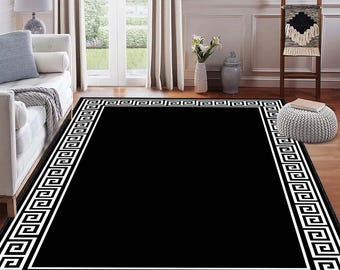 May include: A rectangular black rug with a white Greek key border. The rug is placed on a hardwood floor in a room with a white sofa, a window, and a decorative ladder.