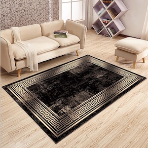 May include: A rectangular black rug with a gold Greek key border. The rug has a distressed appearance with lighter shades of gray and gold. The rug is placed on a light wood floor, with a beige sofa and ottoman in the background.