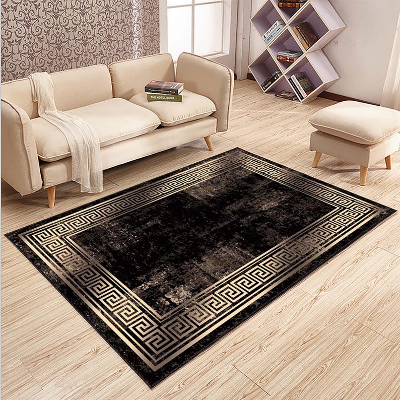 May include: A rectangular black rug with a gold Greek key border. The rug has a distressed appearance with lighter shades of gray and gold. The rug is placed on a light wood floor, with a beige sofa and ottoman in the background.