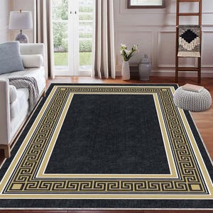 Pode incluir: Um tapete retangular preto com uma borda grega dourada e preta. O tapete está colocado numa sala com um sofá branco, uma escada de madeira e uma porta branca que conduz a um jardim.