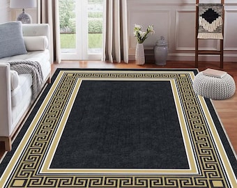 May include: A rectangular black area rug with a gold and black Greek key border. The rug is centered in a room with a white sofa, a wooden ladder, and a white door leading to a garden.
