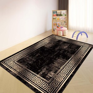 May include: A rectangular black rug with a gold Greek key border. The rug has a distressed appearance with lighter shades of gray and beige. The rug is placed on a light-colored wooden floor in a room with a bookshelf and children's toys.