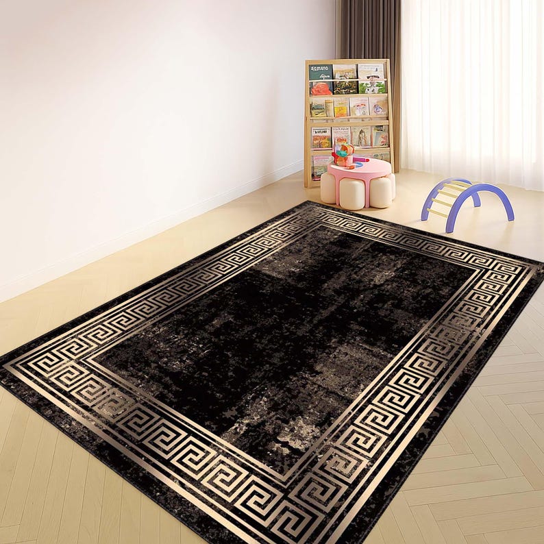 May include: A rectangular black rug with a gold Greek key border. The rug has a distressed appearance with lighter shades of gray and beige. The rug is placed on a light-colored wooden floor in a room with a bookshelf and children's toys.