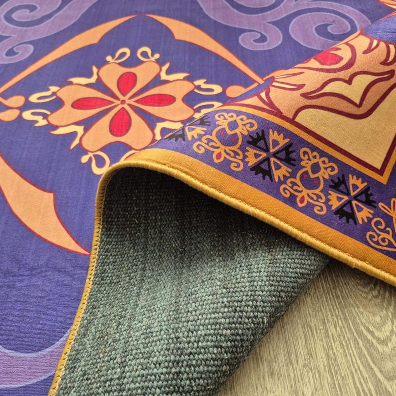 May include: A rolled-up purple rug with an intricate floral and geometric design in shades of orange, red, and gold. The rug's border features a repeating pattern with a gold trim. The underside of the rug is a textured gray fabric.
