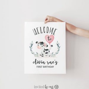 May include: Welcome to Olivia Rae's First Birthday party sign featuring a black and white cow with a pink heart balloon. The sign has a green leafy border.