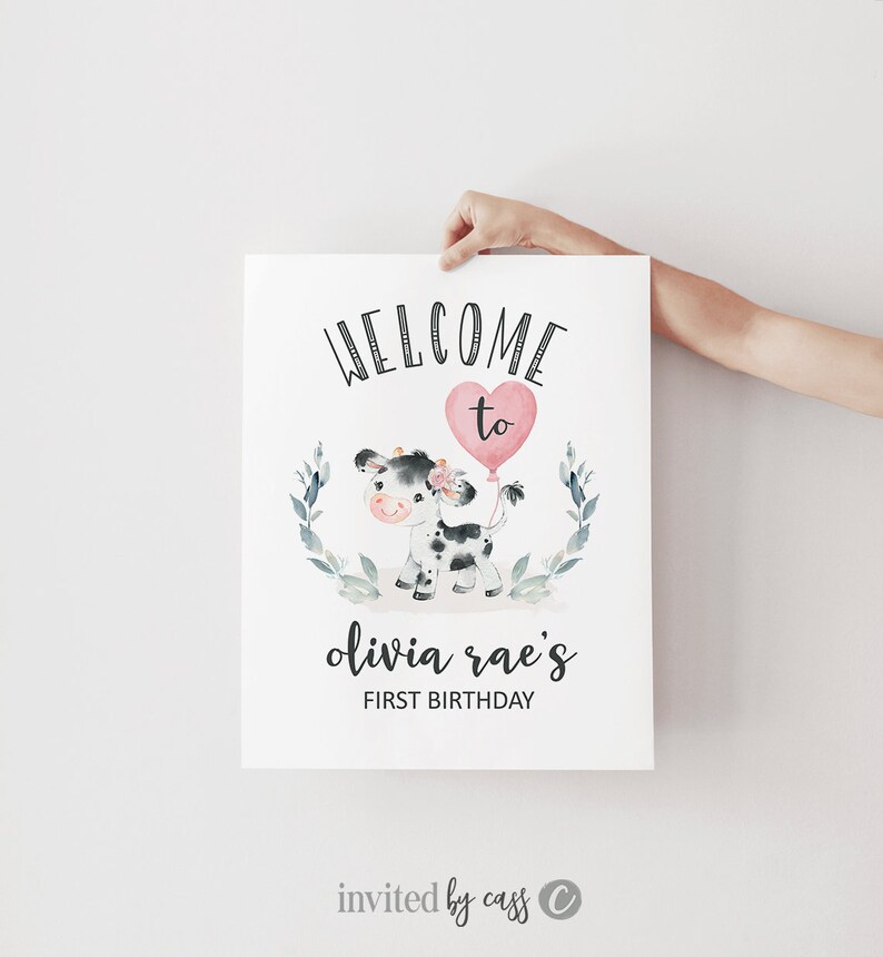 May include: Welcome to Olivia Rae's First Birthday party sign featuring a black and white cow with a pink heart balloon. The sign has a green leafy border.