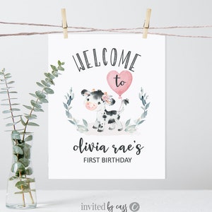May include: Welcome to Olivia Rae's First Birthday sign with a cute cartoon cow holding a pink heart balloon. The sign is decorated with green leaves and flowers.