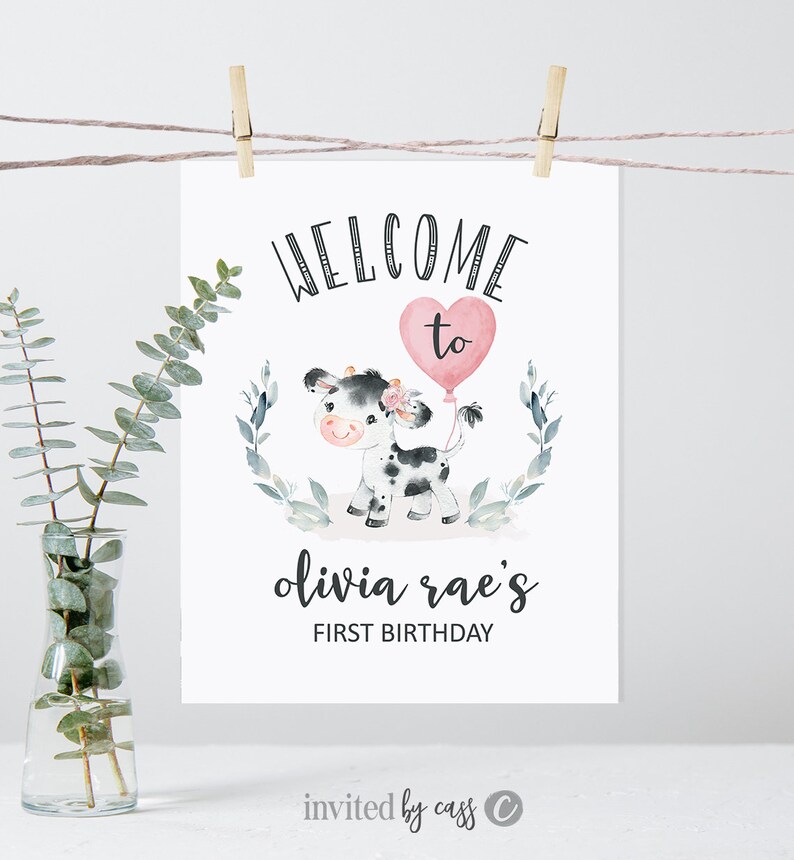 May include: Welcome to Olivia Rae's First Birthday sign with a cute cartoon cow holding a pink heart balloon. The sign is decorated with green leaves and flowers.