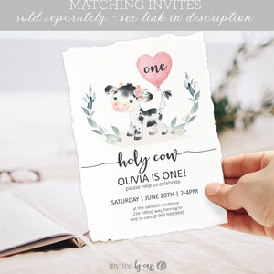 May include: A pink and white cow with a pink heart balloon that says "one" on it. The text "holy cow" is written in cursive. The text "Olivia is one!" is written in black. The text "Please help us celebrate" is written in black. The text "Saturday | June 20th | 2-4pm" is written in black. The text "at the sandhill residence" is written in black. The text "1234 hilltop way, herring tn" is written in black. The text "rsvp to cass @ 999.999.9999" is written in black. The text "invited by cass" is written in black.