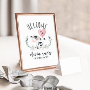 May include: A printable welcome sign for a first birthday party. The sign features a black and white cow with a pink balloon and the text "Welcome to Olivia Rae's First Birthday".