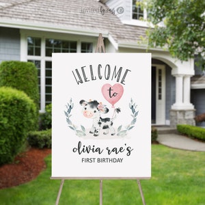 May include: Welcome to Olivia Rae's First Birthday sign with a cute cartoon cow holding a pink heart balloon. The sign is white with black text and a green leafy border.