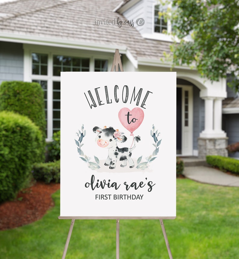 May include: Welcome to Olivia Rae's First Birthday sign with a cute cartoon cow holding a pink heart balloon. The sign is white with black text and a green leafy border.