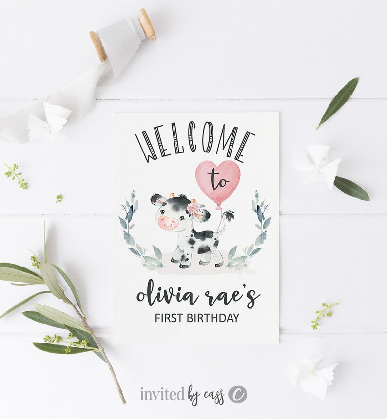 May include: Welcome to Olivia Rae's First Birthday party invitation with a cute cartoon cow holding a pink heart balloon. The invitation has a green leafy border.