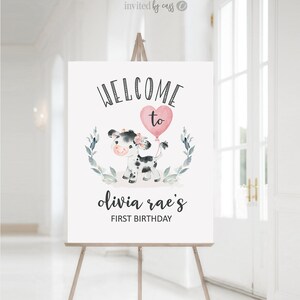 May include: Welcome to Olivia Rae's First Birthday sign with a black and white cow holding a pink heart balloon. The sign is decorated with green leaves.