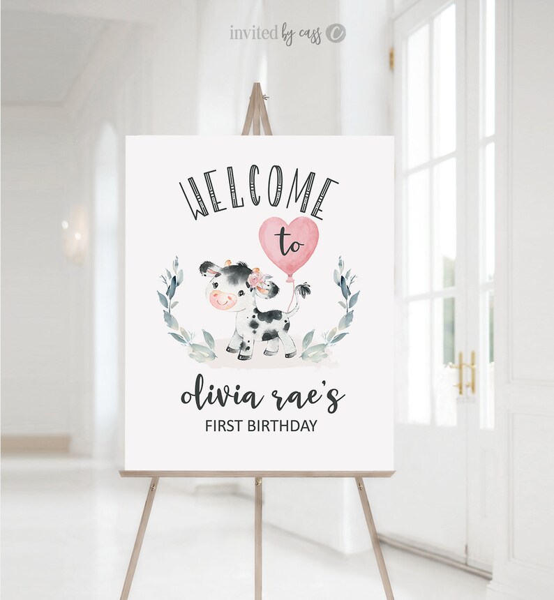 May include: Welcome to Olivia Rae's First Birthday sign with a black and white cow holding a pink heart balloon. The sign is decorated with green leaves.