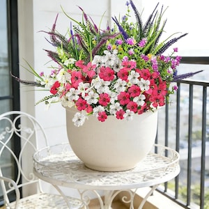 May include: A large white ceramic flower pot overflowing with a vibrant mix of artificial flowers. The arrangement includes red, white, purple, and green foliage, creating a colorful display. The pot sits on a white, ornate metal table, suggesting outdoor decor.