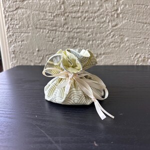 May include: A small, round fabric pouch with a drawstring closure. The pouch is made of a light green and white patterned fabric. The drawstring is a cream-colored ribbon. The pouch is sitting on a dark surface.