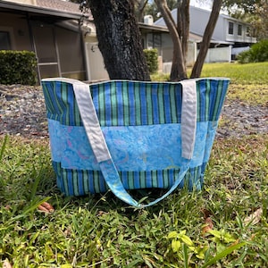 May include: A blue and green striped tote bag with white straps. The bag features a central panel with a blue and pink patterned design. The bag is sitting on a bed of green grass.