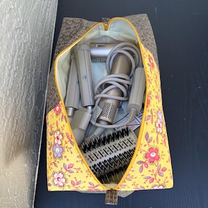 May include: A zippered travel bag with a yellow floral pattern and gray accents, open to reveal hair styling tools. The bag's interior is lined with yellow fabric. The bag contains a hair dryer, curling iron, and various brush attachments.