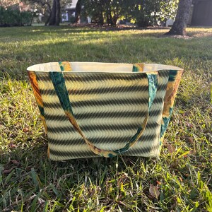 May include: A rectangular tote bag with a wavy pattern in shades of green and yellow. The bag has green straps and a cream-colored interior. The bag is sitting on a grassy surface.