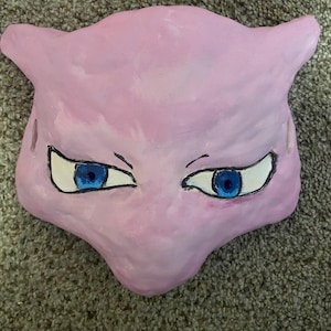 May include: A handmade, pink mask resembling the Pokémon character Mew. The mask features large blue eyes with black outlines and white highlights. The mask has pointed ears and a defined snout, crafted from a soft, textured material.
