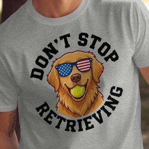 Peut inclure: T-shirt gris avec un dessin de Golden Retriever portant des lunettes de soleil aux couleurs du drapeau américain et tenant une balle de tennis. Le texte "DON'T STOP RETRIEVING" est imprimé autour du chien.