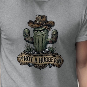 May include: A light gray crew neck t-shirt featuring a graphic of a green cactus wearing a brown cowboy hat. The cactus has a stern facial expression and is positioned above a decorative banner with the text NOT A HUGGER in black lettering.