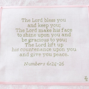 May include: A rectangular needlepoint canvas featuring the text The Lord bless you and keep you; The Lord make his face to shine upon you and be gracious to you; The Lord lift up his countenance upon you and give you peace. Numbers 6:24-26 in olive green thread on a white grid, bordered by a light pink fabric edge.