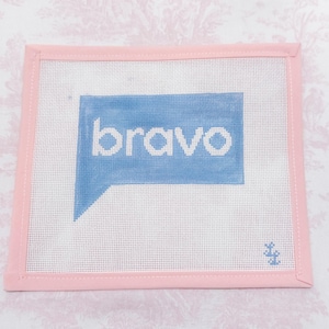 May include: A square needlepoint canvas featuring a light blue speech bubble with the word bravo written in white lowercase letters. The canvas has a light pink fabric border stitched around the edges and a small blue signature mark in the corner.