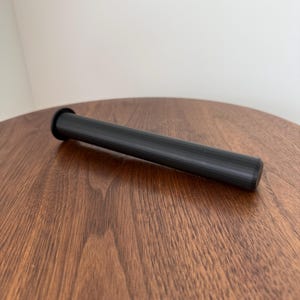 May include: A black, cylindrical object with a flared end, resting on a wooden surface. The object appears to be a container or a part of a larger item. The wood grain of the surface is visible.