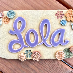 May include: A rectangular ceramic plaque with the name "Lola" in lavender script. The plaque is decorated with colorful ceramic flowers and leaves in shades of orange, blue, and green. The background is a light beige.