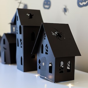 May include: A collection of black paper houses featuring intricate cutouts of spiders, ghosts, and arched windows. These structures are arranged in a row, creating a dark, architectural display with sharp, angular rooflines and detailed silhouettes.