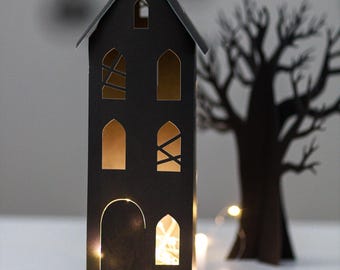 May include: A black paper house with arched windows and a pitched roof. The windows are cut out, revealing a warm glow from the lights inside. A paper tree silhouette stands nearby, creating a spooky scene.