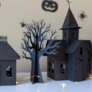 May include: A set of black paper cutouts featuring a house with a fence, a bare tree, a church with a steeple, and a tall building. The structures have arched windows and cut-out details like a cauldron and bat, illuminated by a thin string of lights.