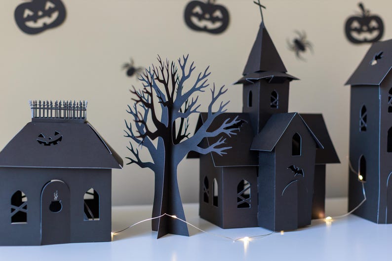 May include: A set of black paper cutouts featuring a house with a fence, a bare tree, a church with a steeple, and a tall building. The structures have arched windows and cut-out details like a cauldron and bat, illuminated by a thin string of lights.