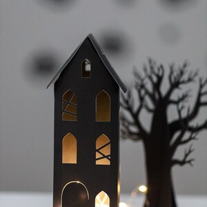 May include: A tall, black paper house structure with arched windows and a door, illuminated from within by warm string lights. Beside the house stands a black paper tree silhouette, creating a dark, layered scene with glowing light accents.