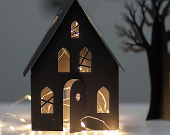May include: A black paper house with cut-out windows and a door, lit by warm white fairy lights. The house features a pitched roof and a gothic design. A paper tree silhouette is in the background, creating a Halloween or festive theme.