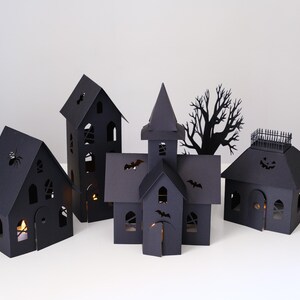 May include: A set of four black paper structures including a tall house, a church-like building with a steeple, a smaller house with a roof railing, and a house with a spider cutout, all featuring illuminated windows and various bat silhouettes.