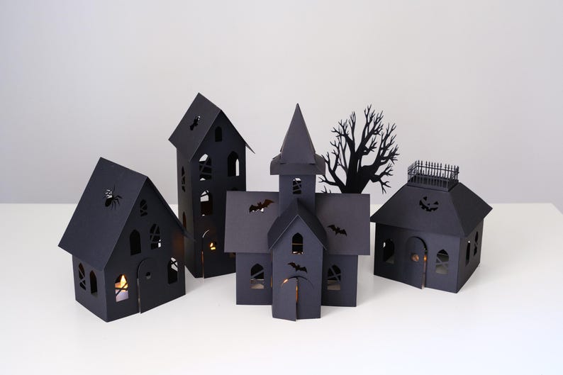 May include: A set of four black paper structures including a tall house, a church-like building with a steeple, a smaller house with a roof railing, and a house with a spider cutout, all featuring illuminated windows and various bat silhouettes.