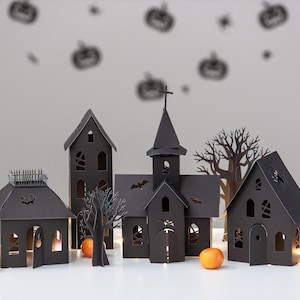 May include: A set of black paper cut-out structures including a tall church with a steeple, two houses, and two bare trees. The buildings feature intricate cut-out windows, doors, and bat silhouettes. Small orange fruits are placed in the foreground.