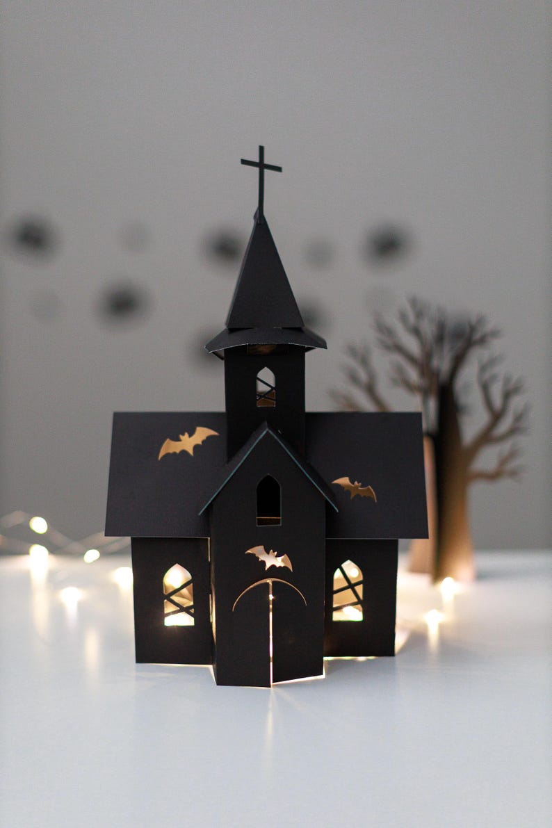 May include: A black paper church structure featuring a tall steeple with a cross on top, arched windows, and cut-out bat silhouettes on the roof and front facade. The structure is illuminated from within, casting a warm glow through the openings.