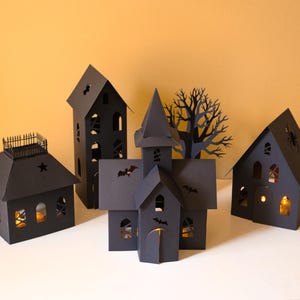 May include: A set of four black paper structures including a tall house, a church with a steeple, a house with a roof railing, and a house with a spider cutout, all featuring intricate window and bat silhouettes with warm internal lighting.