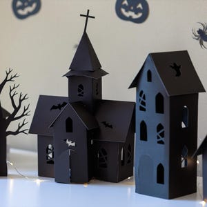 May include: A collection of black paper cutouts featuring a church with a steeple and cross, a tall house, and a bare tree. The structures have intricate cutouts of windows, doors, bats, and ghosts, illuminated by a thin string of warm white lights.