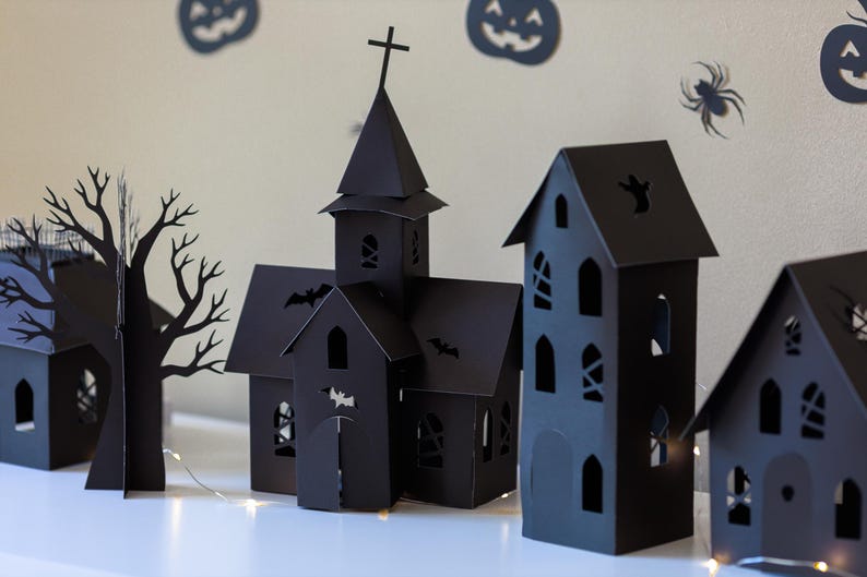 May include: A collection of black paper cutouts featuring a church with a steeple and cross, a tall house, and a bare tree. The structures have intricate cutouts of windows, doors, bats, and ghosts, illuminated by a thin string of warm white lights.