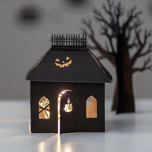 May include: A black paper house decoration featuring a cutout jack-o'-lantern on the roof, a fence along the top ridge, and arched windows. The structure is illuminated from within by warm string lights, casting a glow through the cutouts.