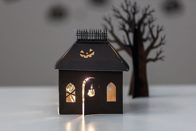 May include: A black paper house decoration featuring a cutout jack-o'-lantern on the roof, a fence along the top ridge, and arched windows. The structure is illuminated from within by warm string lights, casting a glow through the cutouts.