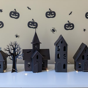 May include: A set of four black paper structures including a house with a roof fence, a bare tree, a church with a steeple, and two tall houses, all featuring cut-out windows and doors, illuminated by a thin string of small white LED lights.